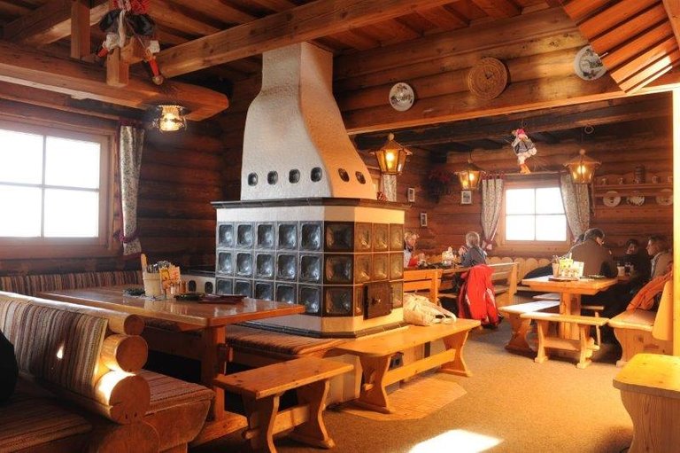 Gemütlicher Gastraum mit Holzwänden, Polsterbänken, Holztischen und Kachelofen, warm beleuchtet. | © Weitmoosalm